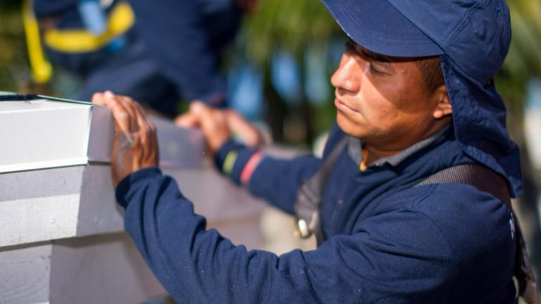 Worker in a blue hooded jacket tightening a joint on a wooden beam outdoors.