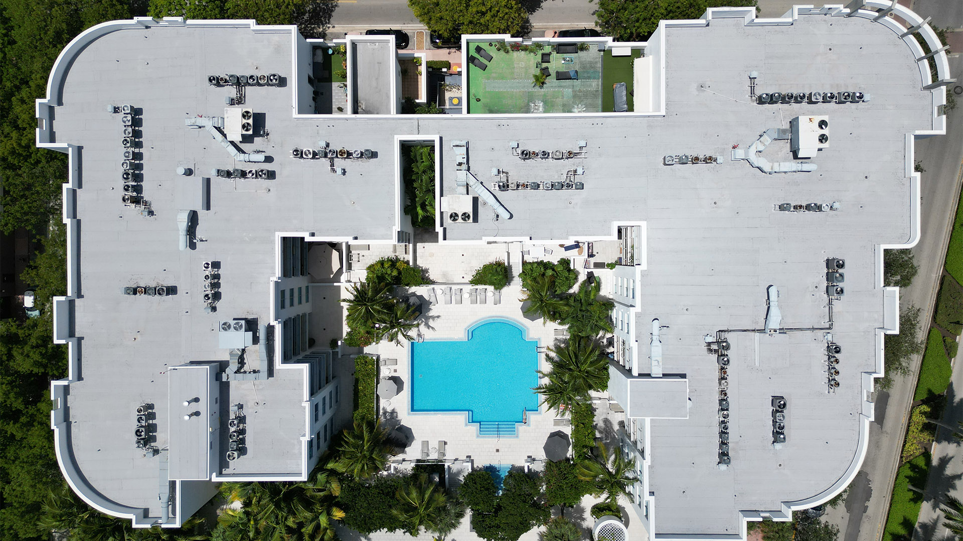 Aerial view of a large white building with a central blue swimming pool, palm trees, and rooftop HVAC units around the edges.