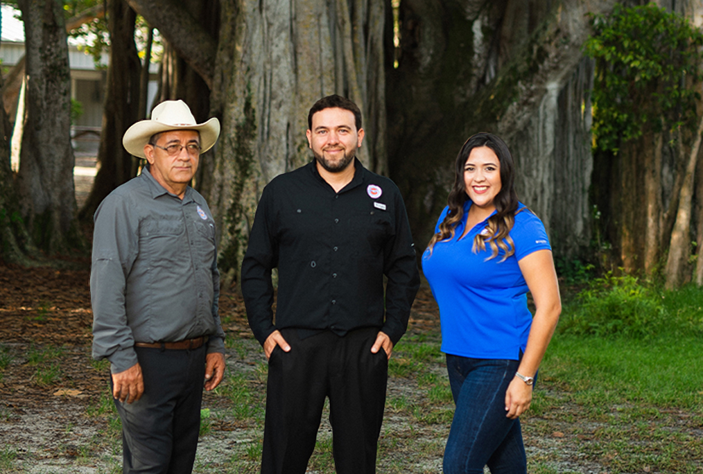 Three adults posing together outdoors in a park: a man in a gray shirt and cowboy hat, a man in a black shirt, and a woman in a blue polo.