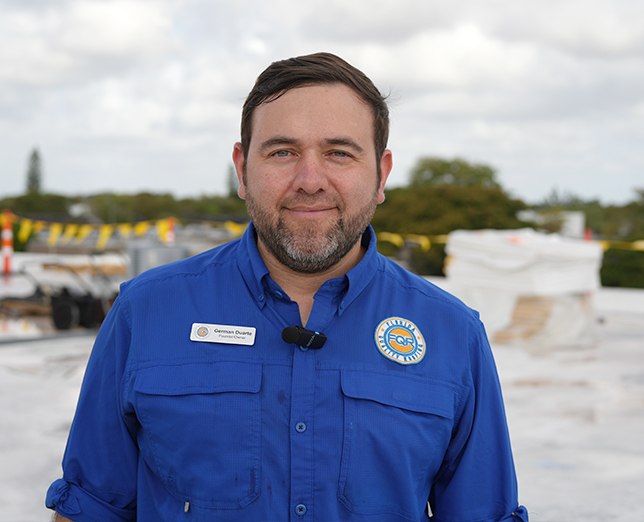 Portrait of a man in a blue uniform with a name badge and microphone, standing on a rooftop construction site with equipment in the background.