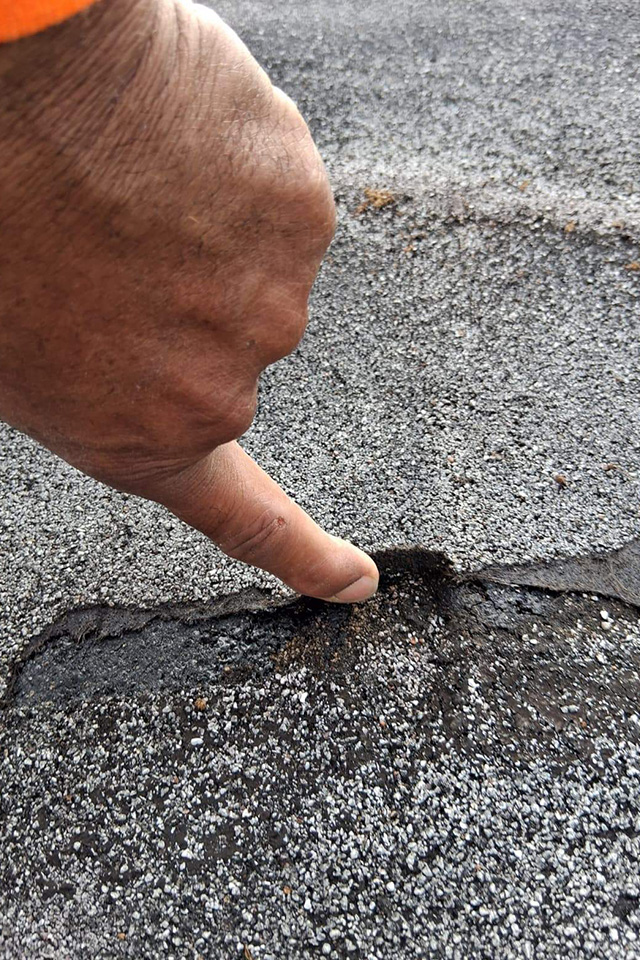 Close-up of a hand pointing at a small crack in coarse asphalt pavement.