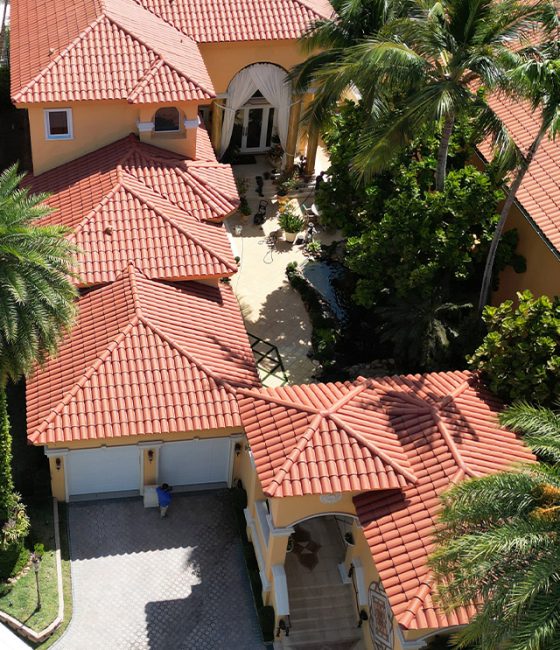 Aerial view of a yellow Mediterranean villa with red tile roofs, palm trees, and a paved courtyard.