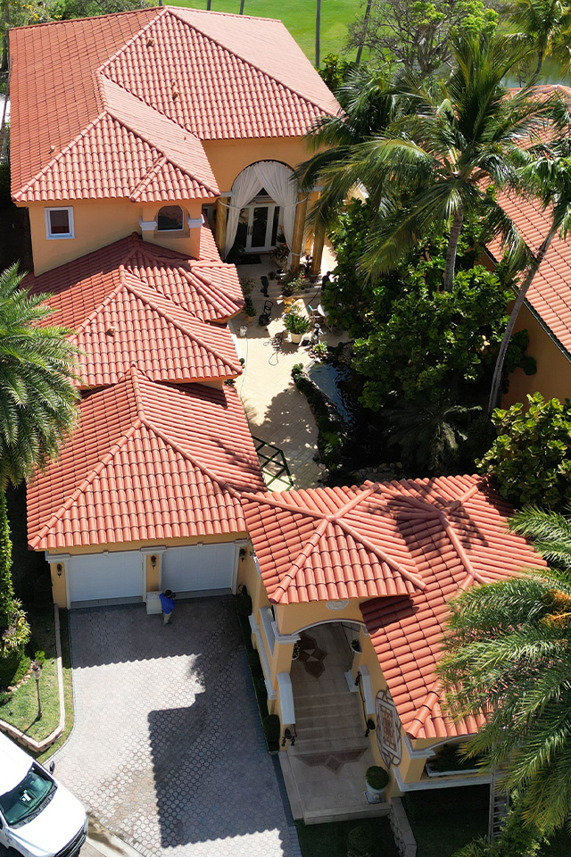 Aerial view of a yellow Mediterranean villa with red tile roofs, palm trees, and a paved courtyard.