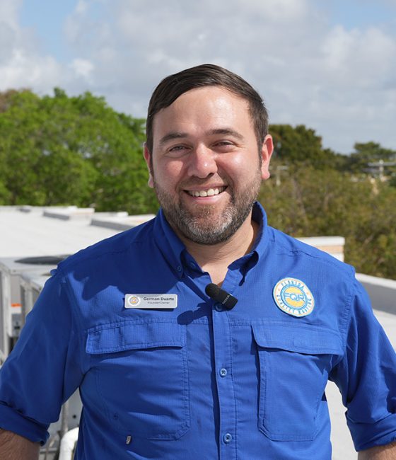Smiling man in a blue work shirt with a name badge on a rooftop, blue sky in the background.