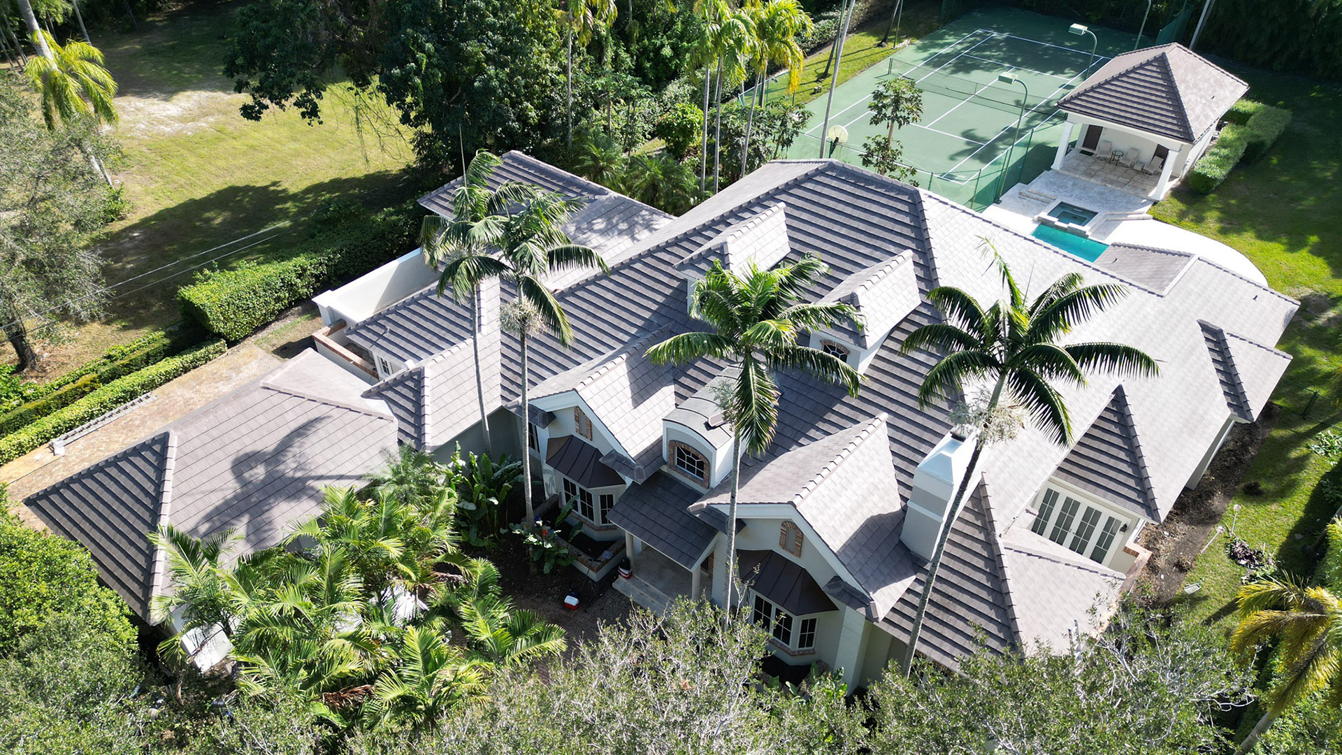 Aerial view of a large modern house with gray tiled roofs, surrounded by palm trees and a tennis court in the background.